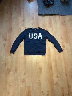 Mörkblå Tommy hilfiger  med USA-tryck - Cool mörkblå Tommy hilfiger med stort vitt USA-tryck på bröstet. Tröjan har rund halsringning och långa ärmar. Perfekt för dig som gillar sportig och avslappnad stil.