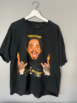 Svart Post Malone t-shirt - Cool svart t-shirt med stort tryck av Post Malone på framsidan, där han har en glorie och eldflammor på fingrarna. Baksidan har ett gult Post Malone-tryck med taggtråd. Oversized passform och tillverkad i mjuk bomull.