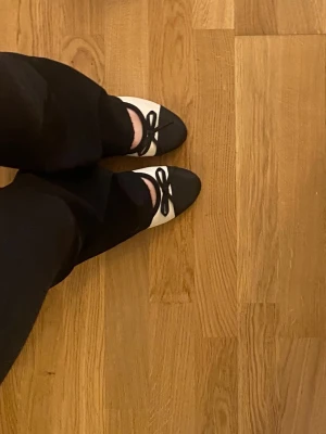 Svartvita ballerinaskor med rosett - Säljer ett par ballerinaskor i klassisk modell med svart tåparti och rosett framtill. Skorna är vita med svarta detaljer och har rundad tå. Perfekta för dig som gillar en stilren och tidlös look.