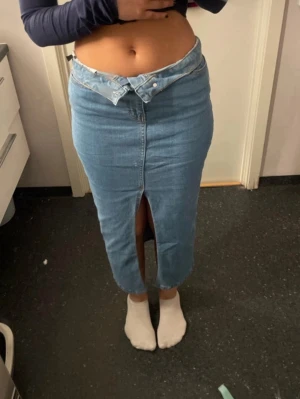 Ljusblå jeanskjol med slits framtill - Trendig ljusblå jeanskjol i lång modell med hög midja och cool asymmetrisk knappstängning. Kjolen har en slits framtill och bakficka, perfekt för en avslappnad men snygg look. Tillverkad i klassiskt denimtyg.