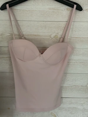 Ljusrosa bustier linnetopp XXS - Säljer en ljusrosa bustier linnetopp med justerbara smala axelband och lätt vadderade kupor. Toppen har en mjuk och stretchig känsla, perfekt för att styla med jeans eller kjol. Enkel och stilren design.