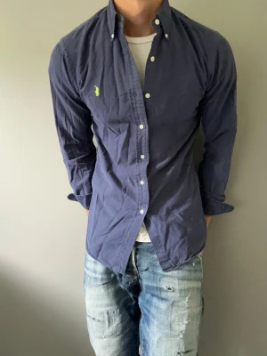 Marinblå skjorta från Ralph Lauren - Säljer en marinblå skjorta från Ralph Lauren med klassisk button-down krage och vit knappstängning. Skjortan har en grön broderad logga på bröstet och är tillverkad i bomull. Perfekt till jeans för en avslappnad men snygg look.