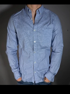 Blå rutig skjorta med knappar - Snygg blå skjorta med diskret rutigt mönster och klassisk krage. Skjortan har lång ärm, bröstficka och vita knappar framtill. Perfekt för en clean och stilren look. Tillverkad i bomull för skön känsla.