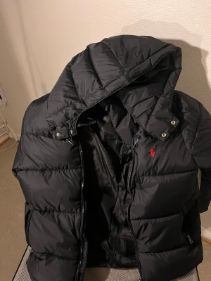 Svart pufferjacka från Polo Ralph Lauren - Säljer en svart pufferjacka från Polo Ralph Lauren med röd broderad logga på bröstet. Jackan har huva, dragkedja framtill och flera fickor med dragkedja. Perfekt för kalla dagar och riktigt skön att bära. Klassisk och stilren design. Fick den som en födelsedag present, använder ej den nu.