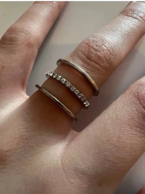 Trebandsring i silver med stenar - Säljer en snygg trebandsring i silverfärg där mittenbandet är dekorerat med gnistrande stenar. Ringen har en modern och stilren design som sticker ut på fingret. Perfekt för dig som gillar minimalistiska smycken med lite extra bling.