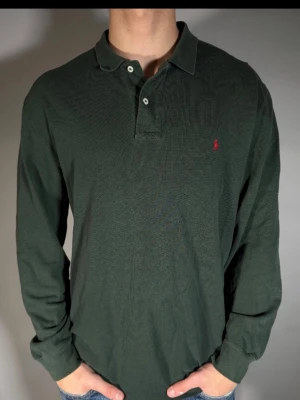 Mörkgrön långärmad skjorta Polo Ralph Lauren - Klassisk mörkgrön långärmad skjorta från Polo Ralph Lauren med krage och knappar vid halsen. Skjortan har en liten röd broderad logga på bröstet och är gjord i mjuk bomull. Perfekt för en avslappnad men ändå stilren look.