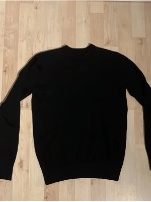 John henric tröja - En enkel svart tröja med rund halsringning och långa ärmar. Tröjan har en klassisk passform och ribbade muddar vid ärmslut och nederkant. Perfekt att matcha med jeans eller över en skjorta för en clean look.