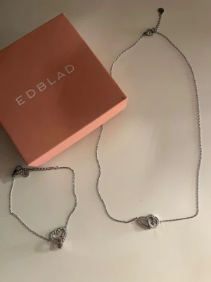 Silverhalsband med hjärthänge från Edblad - Snyggt silverfärgat halsband från Edblad med ett hjärthänge. Kedjan är tunn och gjort av rostfritt stål. Matchande armband medföljer. Den är oandvänd och kommer i original ask.