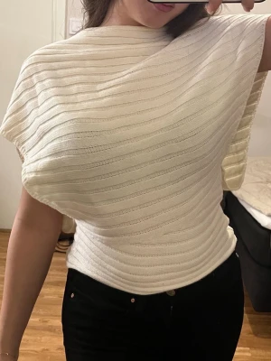 Vit ribbad topp med asymmetrisk ärm - Säljer en vit ribbad topp med unik asymmetrisk design och breda, korta ärmar. Toppen har en kroppsnära passform i midjan och är tillverkad i ett mjukt, stretchigt material. Perfekt för dig som gillar stilrena och moderna plagg. Använt en gång endast