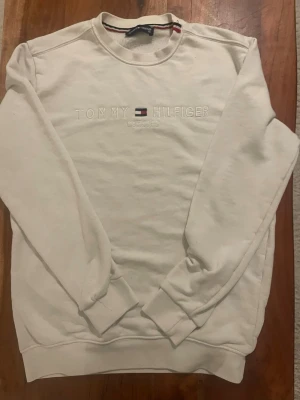 Vit sweatshirt från Tommy Hilfiger - Snygg vit sweatshirt från Tommy Hilfiger med broderad logga och texten 'EST. 1985' på bröstet. Tröjan har rund halsringning, ribbade muddar och klassisk passform. Perfekt för en clean och stilren look.