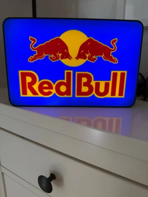 Red Bull ljusskylt i plast - Cool Red Bull ljusskylt med klassisk logga i rött och gult mot blå bakgrund. Skylten är rektangulär och gjord i plast, perfekt som dekoration till rummet eller gaminghörnan. Den har en sladd för att kopplas in och lyser upp med starka färger.