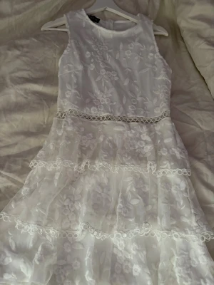 Vit spetsklänning med volanger - Säljer en superfin vit klänning med blommig spets och volanger. Klänningen är ärmlös, har rund halsringning och flera lager med spetsdetaljer. Perfekt för sommarens alla tillfällen! 