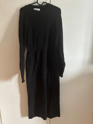 Svart lång stickad klänning AWARE - Lång svart stickad klänning från AWARE. Klänningen har en enkel, rak passform och är perfekt för dig som gillar en stilren look. Materialet är mjukt och ribbat vilket ger en snygg struktur. Med slits baktill 