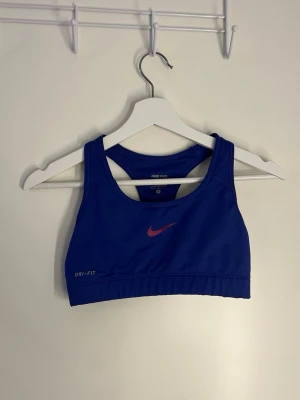 Blå sport-bh från Nike Dri-FIT - Säljer en blå sport-bh från Nike med Dri-FIT-material som transporterar bort fukt. Bh:n har brottarrygg och en röd Nike-logga framtill. 