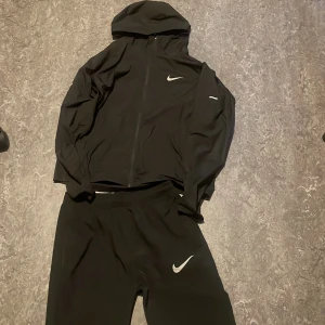 Svart Nike träningsset med huva - Snyggt svart träningsset från Nike med matchande jacka och byxor. Jackan har huva och dragkedja framtill samt Nike-logga på bröstet. Byxorna har resår i midjan och Nike-logga på låret. Perfekt för träning eller chill.