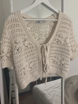 Beige virkad kort kofta från Zara - Säljer en beige, virkad kort kofta från Zara med knytning framtill. Koftan har ett öppet hålmönster och virkade blommor som detaljer. Perfekt att slänga över ett linne eller klänning. Tillverkad i mjukt bomullsmaterial.