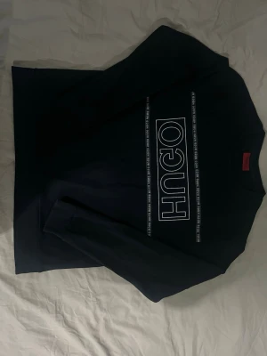 Svart sweatshirt från Hugo - Svart sweatshirt från Hugo med stort vitt logotryck på bröstet och tunna vita linjer med upprepat Hugo-tryck. Klassisk rund hals och långa ärmar. Perfekt för en clean och modern streetwear-look.