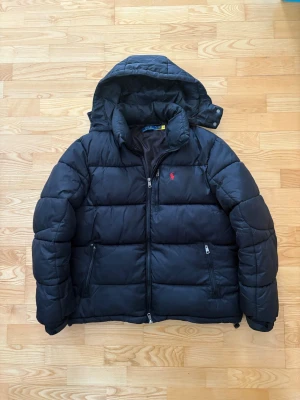  Polo Ralph Lauren jacka - Säljer en svart pufferjacka från Polo Ralph Lauren med huva och dragkedja framtill. Jackan har två snedställda fickor med dragkedja och den klassiska röda loggan broderad på bröstet. Perfekt för kalla vinterdagar och riktigt skön att bära.