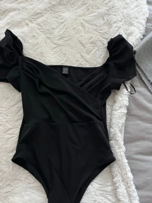 Svart body med puffärmar  - Trendig svart body med snygga puffärmar och omlott-detalj framtill. Tillverkad i ett mjukt och stretchigt material som sitter bekvämt mot kroppen. Perfekt för dig som vill ha en stilren och modern look.