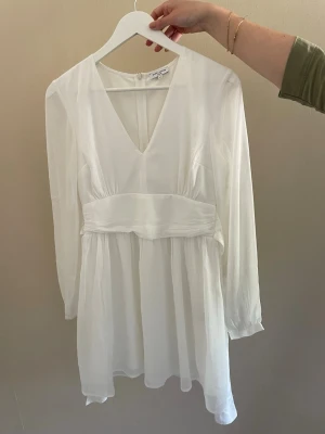 Vit kort omlottklänning med v-ringning - Superfin vit kort klänning från Bubbleroom med v-ringning och markerad midja. Klänningen har långa ärmar och är tillverkad i ett lätt, flowy material som ger en luftig känsla. Perfekt för sommarens alla tillfällen!