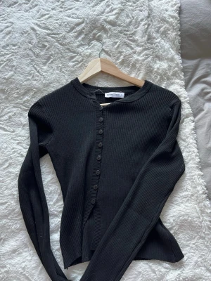 Svart ribbad kofta från Crazy Dizzy - Snygg svart ribbad kofta från Crazy Dizzy med rund halsringning och knappar hela vägen framtill. Långärmad modell i mjukt, stretchigt material som sitter tajt och ger en clean look. Perfekt att styla med jeans eller kjol.