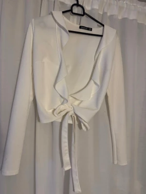 Vit omlottopp med volang från Boohoo - Snygg vit omlottopp från Boohoo med långa ärmar och volangkant längs den djupa v-ringningen. Toppen knyts framtill för en trendig look och har en croppad passform. Perfekt att styla med höga jeans eller kjol.
