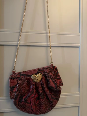 Röd axelväska med ormmönster - Cool handväska i rött och svart ormmönstrat material med en glansig finish. Väskan har en guldfärgad kedjerem och ett hjärtformat guldfärgat spänne framtill. Unik form med veckad design som ger extra attityd.