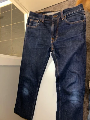 Mörkblå jeans från Nudie Jeans - Säljer ett par mörkblå jeans från Nudie Jeans med klassisk femficksmodell och kontrastsömmar. Jeansen har raka ben och Nudie Jeans ikoniska broderi på bakfickorna. Perfekta för en avslappnad och stilren look.