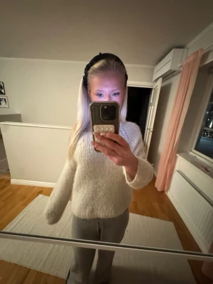 Mjuk vit stickad tröja - En fluffig och mjuk stickad tröja i vit färg med rund hals och långa ärmar. Tröjan har en avslappnad passform och är perfekt för att hålla sig varm under kyliga dagar. Materialet ser ut att vara en blandning av syntet och ull för extra komfort. 