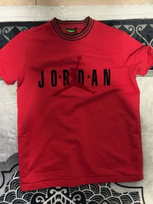 Röd Jordan t-shirt med tryck - Snygg röd t-shirt från Jordan med svart logga och text på bröstet. T-shirten har rund hals med svarta ränder och korta ärmar. Perfekt för dig som gillar sportig stil och streetwear.