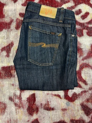 Nudie bootcut ola dry denim - Snygga mörkblå jeans från Nudie med klassisk bootcut passform och kontrastsömmar. Jeansen har en ikonisk bakficka med gul brodyr och patch i midjan. Tillverkade i slitstark denim, perfekta för en avslappnad och trendig look. Storlek 30 32