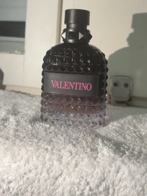 Valentino parfym svart glasflaska - Säljer en lyxig parfym från Valentino i en cool svart glasflaska med nitar och struktur. Flaskan har en gradient från svart till genomskinlig och ett svart lock. Märket står i rosa på etiketten framtill. Perfekt för dig som gillar exklusiva dofter.