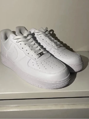 Nike Air Force 1 vita sneakers - Klassiska Nike Air Force 1 sneakers i helvitt läder med perforerad tå, vit sula och matchande vita skosnören. Ikonisk design med Nike swoosh på sidorna och metallbricka med 'AF1' på snörningen. Perfekta för en clean och stilren look.