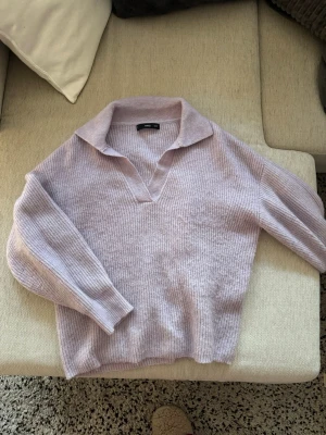 Ljuslila stickad tröja från MANGO - Mysig ljuslila stickad tröja från MANGO med krage och v-ringad hals. Tröjan har långa ärmar och är ribbstickad för extra struktur. Perfekt för att hålla värmen under kyliga dagar och ger en avslappnad men ändå stilren look.