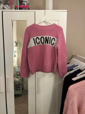 Rosa stickad tröja med text - Säljer en rosa stickad tröja med vit panel och svart texten 'ICONIC' över bröstet. Tröjan har rund halsringning, långa ärmar och ribbade muddar vid ärmslut och nederkant. Perfekt för dig som vill ha en trendig och bekväm look.