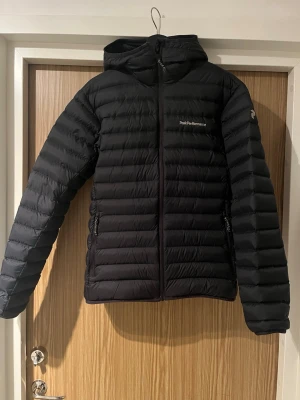 Svart dunjacka från Peak Performance - Snygg svart dunjacka från Peak Performance med huva och dragkedja framtill. Jackan har två sidofickor med dragkedja och diskret logga på bröstet. Lätt och smidig modell med horisontella sömmar som håller dunet på plats. Perfekt för kalla vinterdagar.