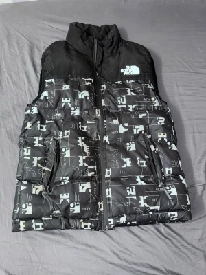 Svart dunväst från The North Face - Cool svart dunväst från The North Face med vit logga på bröstet och ett unikt grafiskt mönster över hela västen. Västen har hög krage, två dragkedjeförsedda fickor och är tillverkad i ett glansigt material. Perfekt för att lägga till lite edge till din outfit.