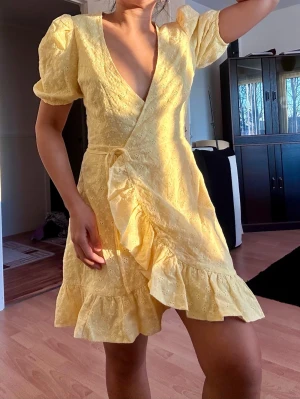 Gul omlottklänning från NA-KD - Söt gul omlottklänning från NA-KD med volangdetaljer och puffärmar. Klänningen har broderade blommor, knyt i midjan och v-ringad hals. Perfekt för sommarens alla tillfällen och ger en fräsch och trendig look.