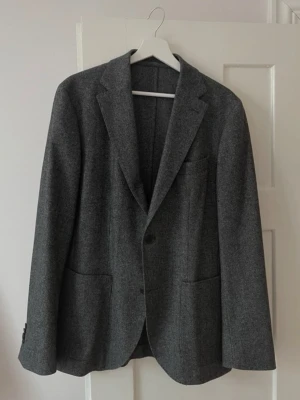 Kavaj från Riley - Stilren mörkgrå kavaj i ull med två knappar framtill, klassiska slag och två stora framfickor. Kavajen har en tidlös design och enkel bröstficka. Perfekt för dig som vill ha en clean och snygg look till vardags eller mer uppklätt.