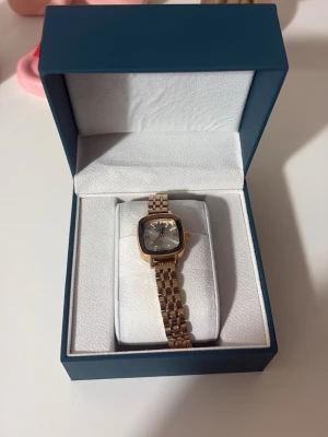 Guldfärgad klocka från MOTF - Stilren klocka från MOTF med fyrkantig urtavla i silver och guldfärgat armband i metall. Urtavlan har romerska siffror och gnistrande stenar som timmarkeringar. Perfekt accessoar för dig som gillar en elegant look.