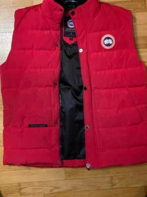 Röd Canada Goose Väst (med deffekter) - Säljer min röda Canada Goose väst som bara står i garderoben och tar plats så vill bli av med den. Den är rätt sliten och har ett par deffekter så som ett hål på högra axeln se på bild 5. Och även ett ihopsydd ficka se bild 6. Och en saknad knapp längst ner på västen se på bild 7.