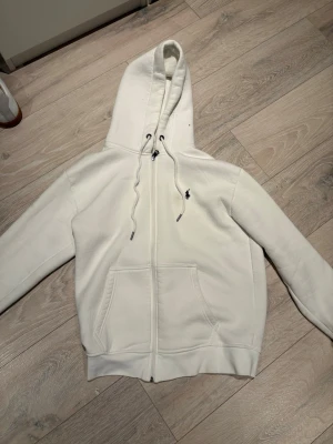 Vit hoodie med dragkedja från Polo Ralph Lauren - Klassisk vit hoodie från Polo Ralph Lauren med dragkedja framtill, två stora fickor och justerbar huva med snören. Liten broderad logga på bröstet. Tillverkad i mjukt bomullsmaterial för en skön och avslappnad look.