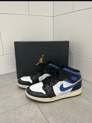 Air Jordan 1 Mid svart/blå/vit - Säljer ett par Air Jordan 1 Mid sneakers i svart, blått och vitt läder. Klassisk siluett med blå Swoosh och Wings-logga på sidan. Perforerad tåbox för ventilation och svart snörning. Perfekta för dig som gillar streetwear och ikoniska sneakers.