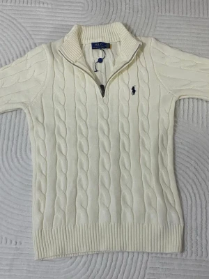 Vit kabelstickad tröja Polo Ralph Lauren - Kabelstickad vit tröja från Polo Ralph Lauren med halv dragkedja och hög krage. Tröjan har det klassiska blå broderade logotypen på bröstet och ribbade muddar vid ärmslut och nederkant. Perfekt för en stilren och tidlös look. Storlek M men passar mer S