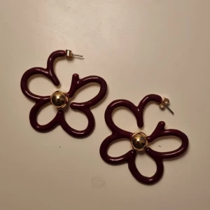 Blommiga örhängen i vinrött och guld - Unika örhängen i form av blommor med vinröda, böjda metallkanter och en guldfärgad pärldetalj i mitten. Perfekta för dig som vill sticka ut med en lekfull och trendig accessoar.