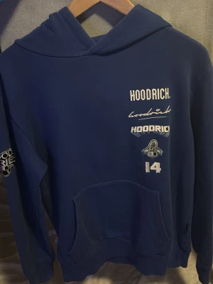 Mörkblå hoodie från Hoodrich - Snygg mörkblå hoodie från Hoodrich med flera vita och broderade loggor och texter på bröstet samt ärmen. Klassisk känguruficka framtill och huva utan dragsko. Perfekt för en streetwear-look och riktigt skön passform.