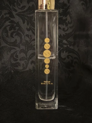 Essens parfym 50 ml glasflaska w114 - Elegant parfym från Essens i en genomskinlig, rektangulär glasflaska med gulddetaljer och guldlock. Flaskan rymmer 50 ml och har en stilren design med guldfärgade cirklar på framsidan. Perfekt för dig som gillar exklusiva dofter.