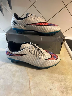 Nike hypervenom phantom 1 - Säljer ett par vita Nike fotbollsskor med blå sula och stora svarta och röda Swoosh-loggor. Skorna har snörning, låg profil och texturerad ovandel för bättre bollkontroll. Perfekta för konstgräs och gräsplaner. Snygg och modern design för dig som vill sticka ut på planen. 