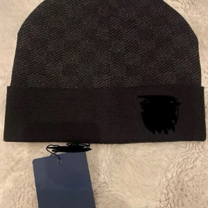 Mörkgrå ribbad mössa med uppvikt kant - Snygg mörkgrå mössa med diskret mönster och bred uppvikt kant. Mössan har en enkel och stilren design, perfekt för att hålla värmen under kalla dagar. Märke syns på kanten. Passar till de flesta outfits.