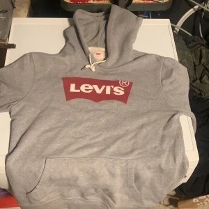 Grå Levi's hoodie med logga - Säljer en grå hoodie från Levi's med stor röd logga på bröstet. Klassisk känguruficka framtill och justerbar huva med snören. Mjuk och skön bomullsblandning, perfekt för chill dagar. Enkel och stilren design som funkar till det mesta.
